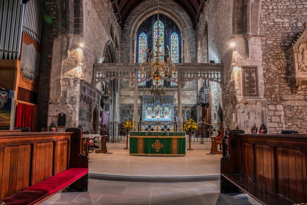 Interior view of St. Mary's Cathedral featuring wooden pews with red cushions, a green altar cloth, vibrant stained glass windows, ornate chandeliers, and floral arrangements flanking the altar.