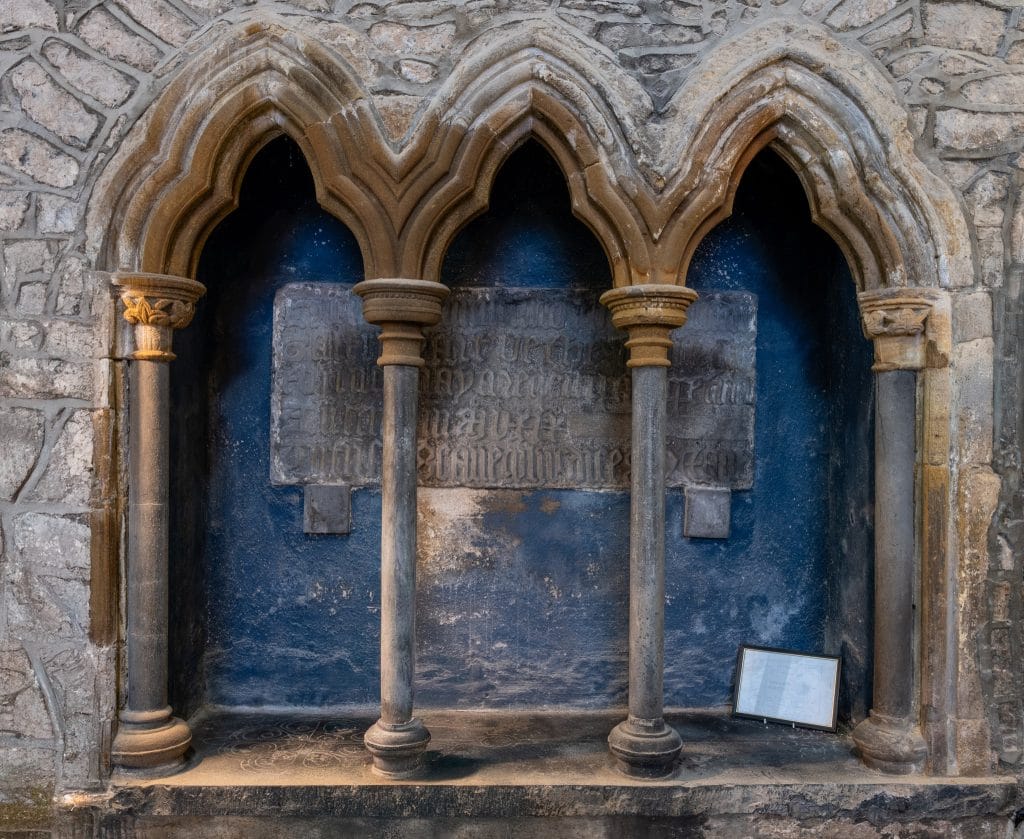 Stone alcove featuring three pointed arch niches separated by four classical columns, with a blue-painted back wall displaying a worn, engraved stone plaque. The surrounding wall is made of rough stone blocks. A framed informational sign rests at the bottom right inside the alcove.