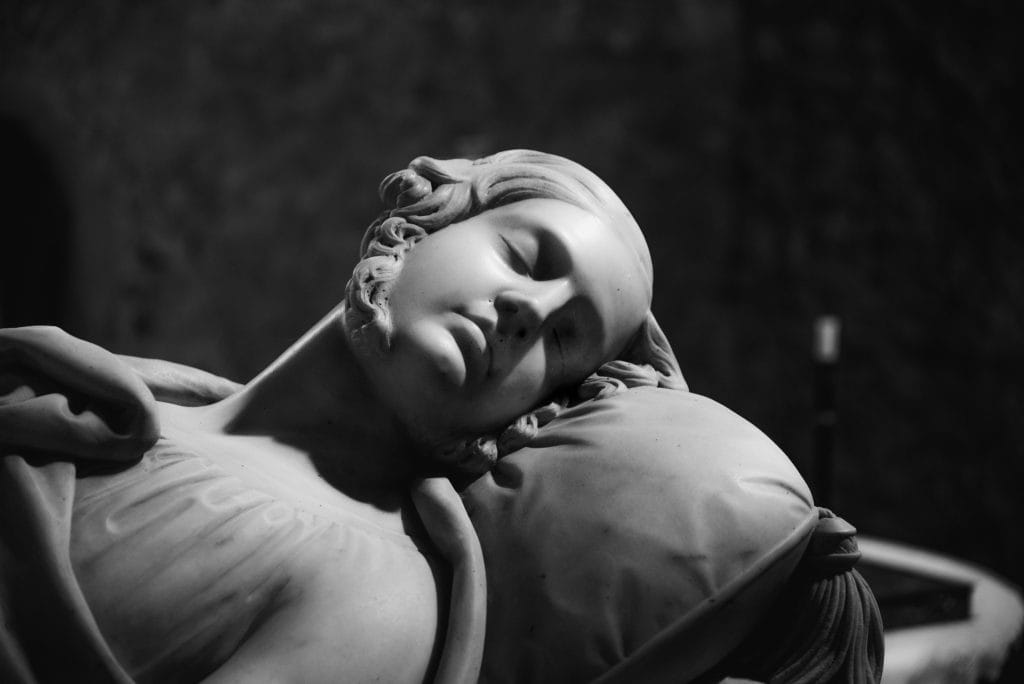 Black and white close-up of a marble statue depicting a reclining figure with closed eyes resting their head on a pillow.