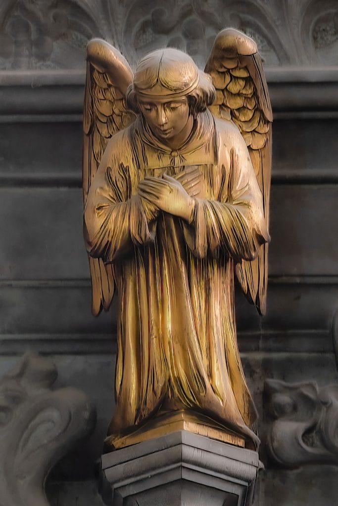 A close-up of a golden angel statue with wings folded behind, hands crossed over its chest, and head bowed, mounted on a stone pedestal against a dark architectural background.