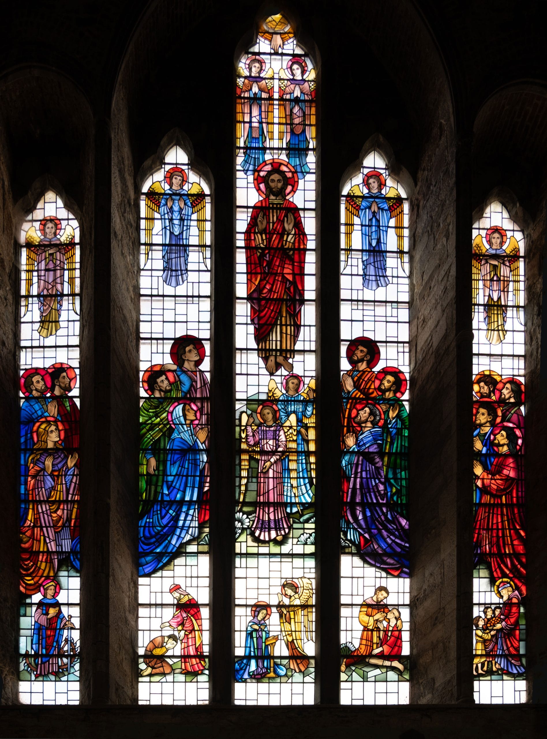 Stained glass window with five tall panels depicting religious figures, including Jesus in red robes at the center, surrounded by angels and disciples in vibrant colors, set within a stone church interior.