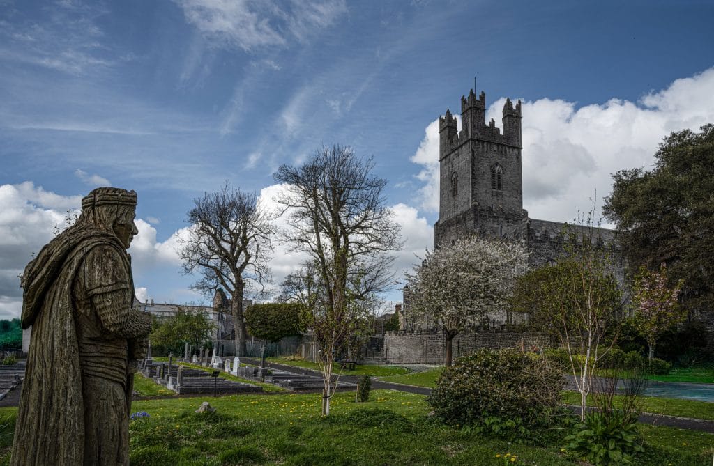 Stone statue of a medieval knight overlooking a churchyard with tombstones, leafless trees, and blooming shrubs under a partly cloudy blue sky, with a tall gray stone church tower in the background.