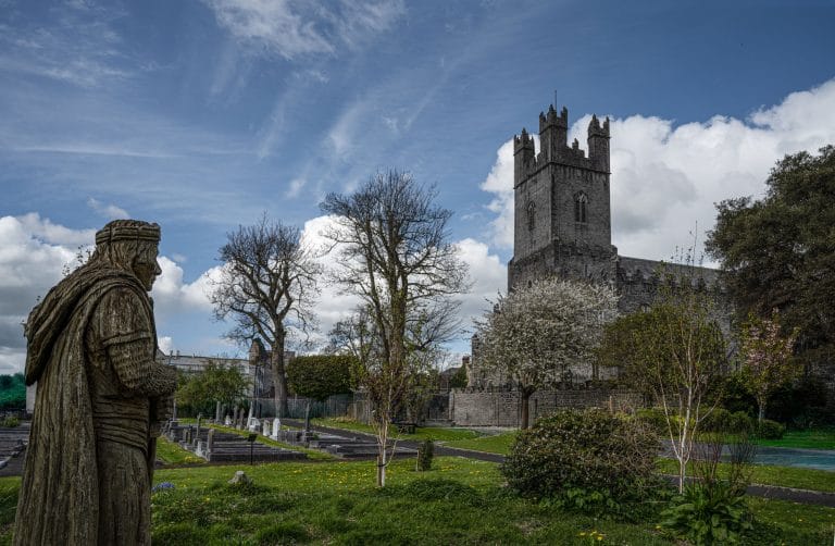 Stone statue of a medieval knight overlooking a churchyard with tombstones, leafless trees, and blooming shrubs under a partly cloudy blue sky, with a tall gray stone church tower in the background.