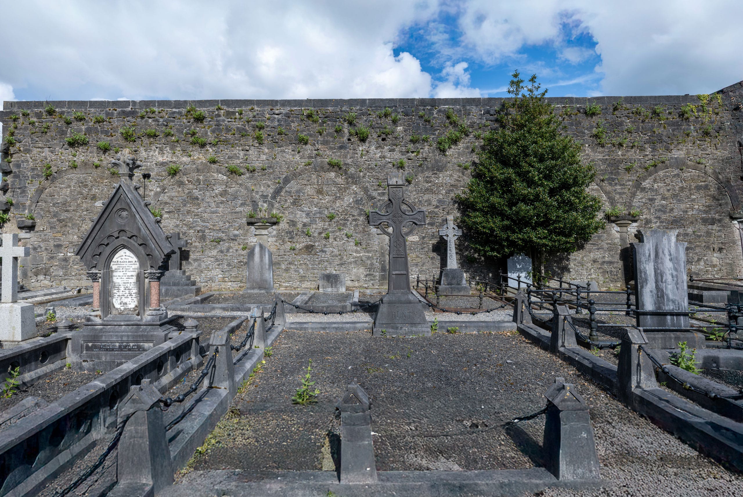 Gravestones and monuments in an old cemetery enclosed by a stone wall with some greenery growing on it, under a partly cloudy blue sky.