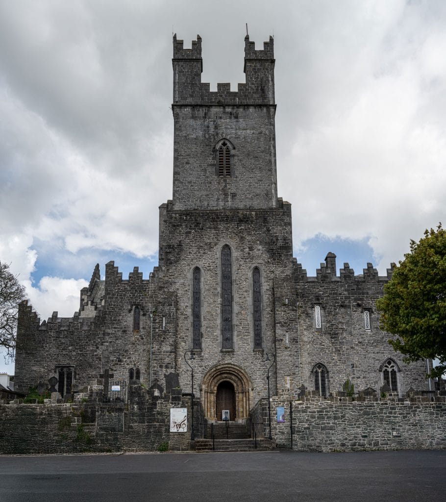 Stone medieval church with a tall rectangular tower featuring battlements, three arched stain-glass windows, an arched wooden entrance door, surrounded by a low stone wall and gravestones under a cloudy sky.