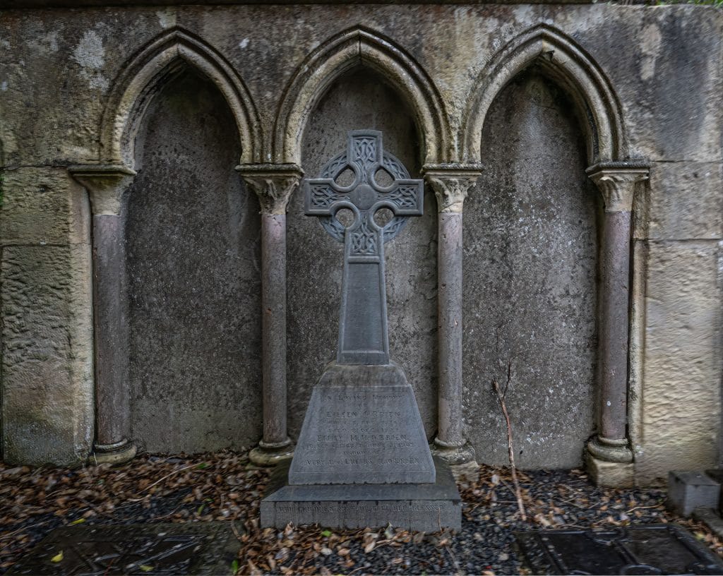 A stone Celtic cross tombstone stands in front of a weathered stone wall with three arched niches supported by stone columns, surrounded by fallen leaves and gravel on the ground.