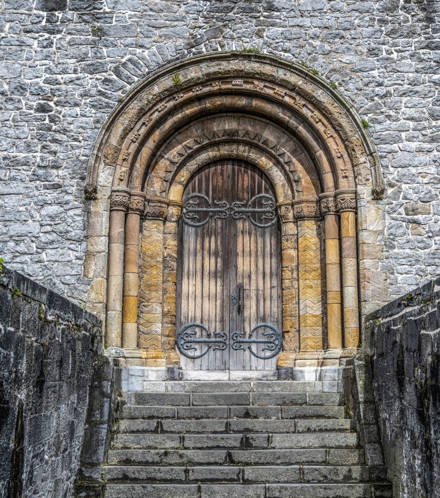 Stone steps lead up to an ornate arched wooden door set in a gray stone wall, featuring intricate stone carvings and decorative metal hinges.