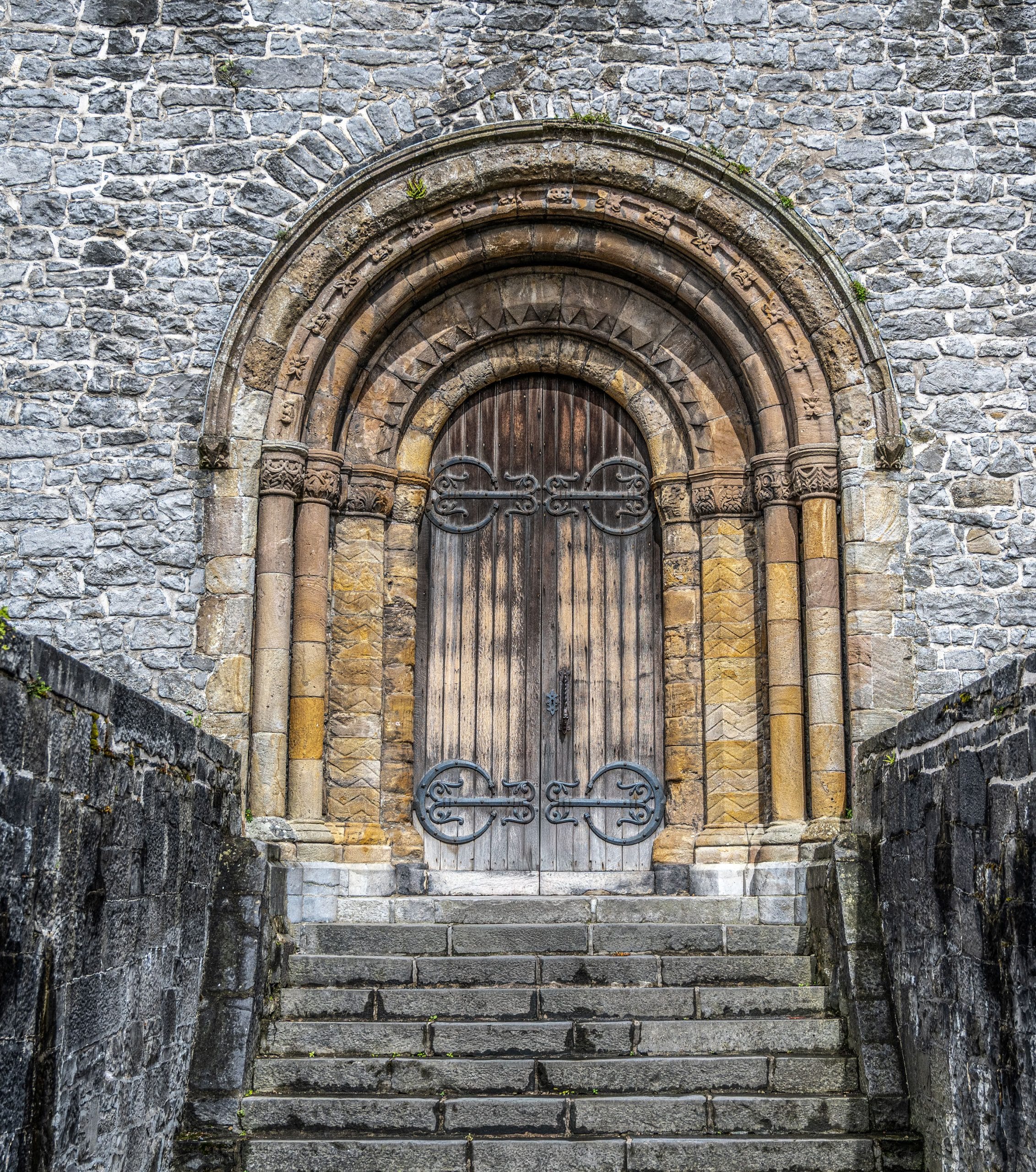 Stone steps lead up to an ornate arched wooden door set in a gray stone wall, featuring intricate stone carvings and decorative metal hinges.