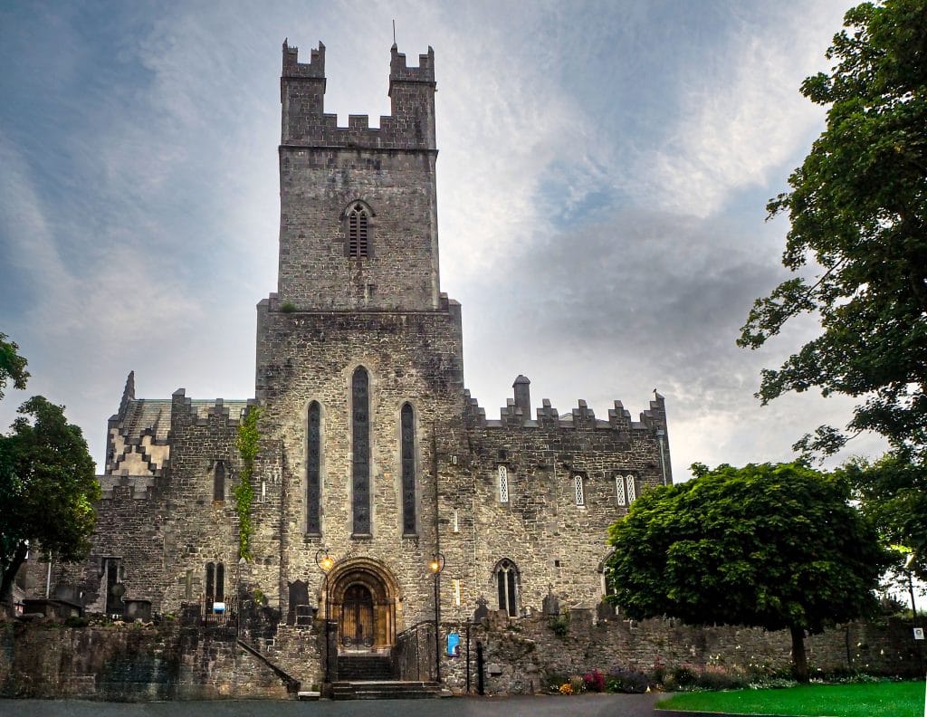 Stone medieval church with a tall square tower, arched door entrance, and narrow vertical windows, surrounded by trees and a low stone wall under a cloudy sky.