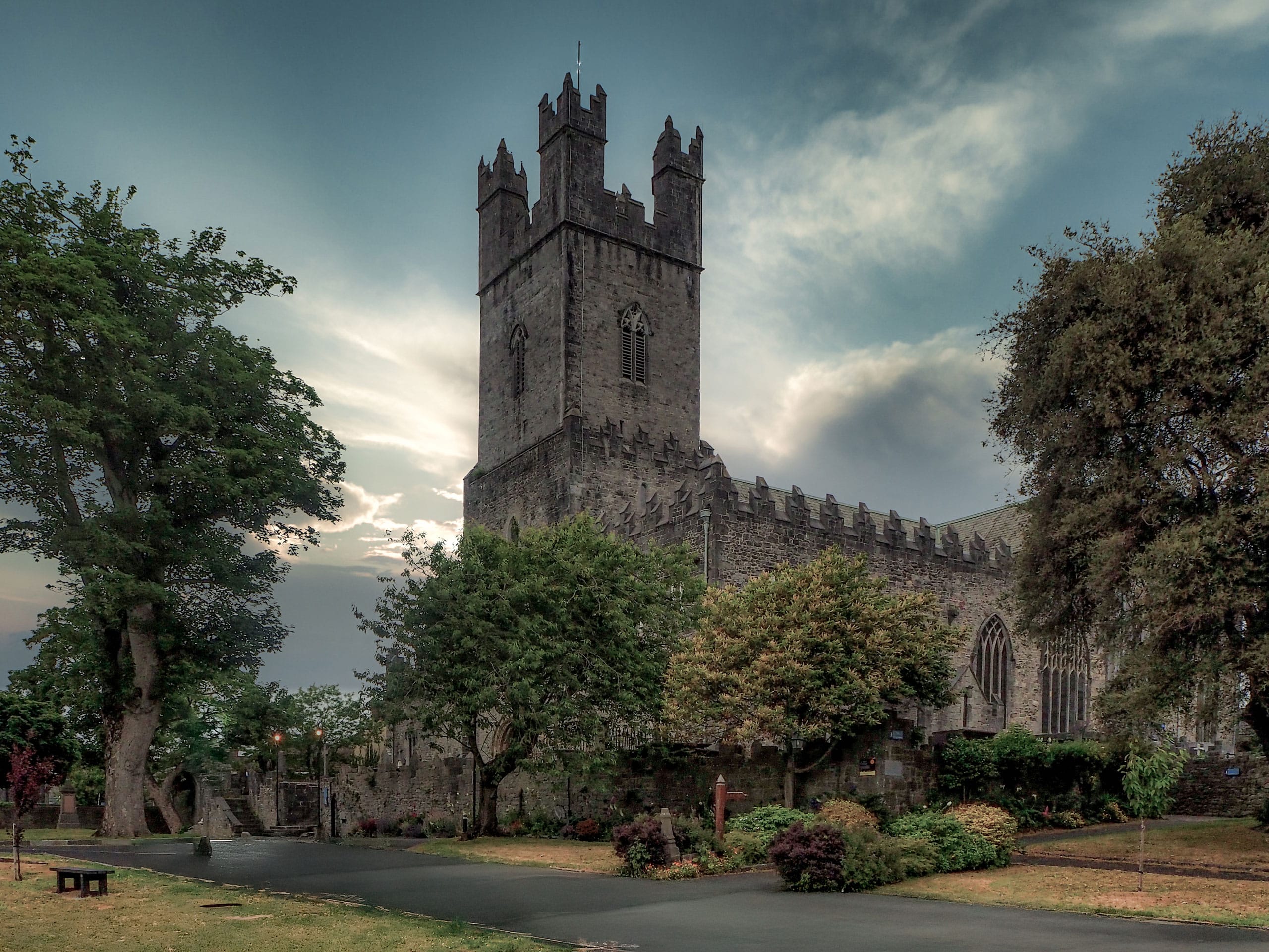 Stone church building with a tall square tower and battlements, surrounded by trees and greenery under a cloudy sky at dusk.