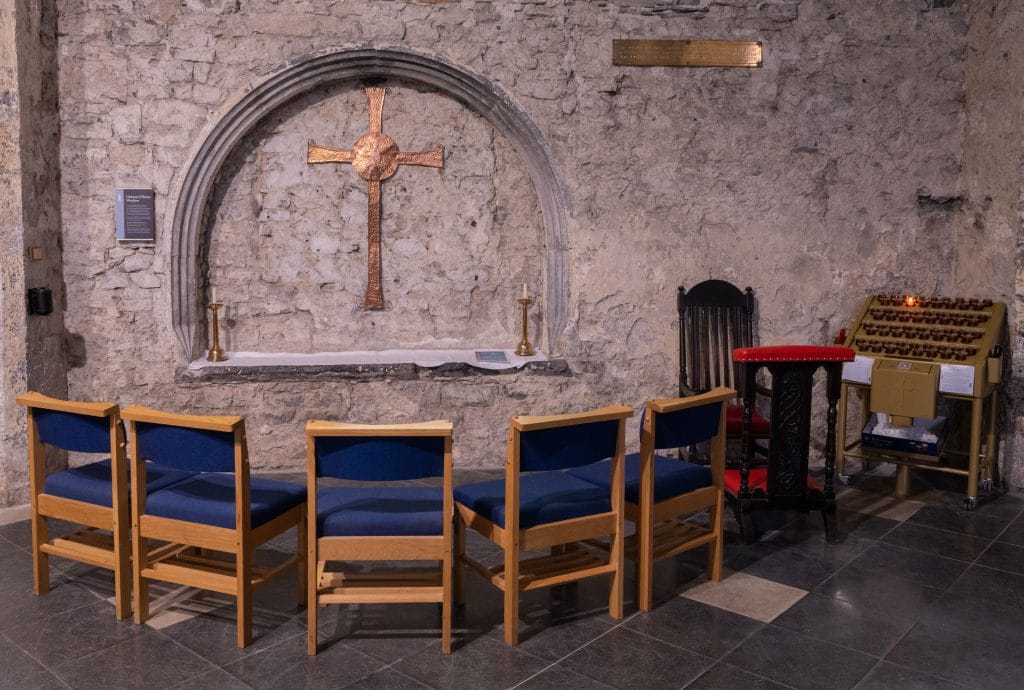 A small stone chapel interior featuring a copper cross mounted inside a stone alcove on a rough stone wall. Below the cross, two candlesticks flank a white cloth-covered ledge. In front, five wooden chairs with blue cushions are arranged in a semicircle. To the right, a wooden kneeling bench with red cushioning and a black chair stand next to a votive candle holder rack with one candle lit. The floor is tiled in large gray and cream squares.