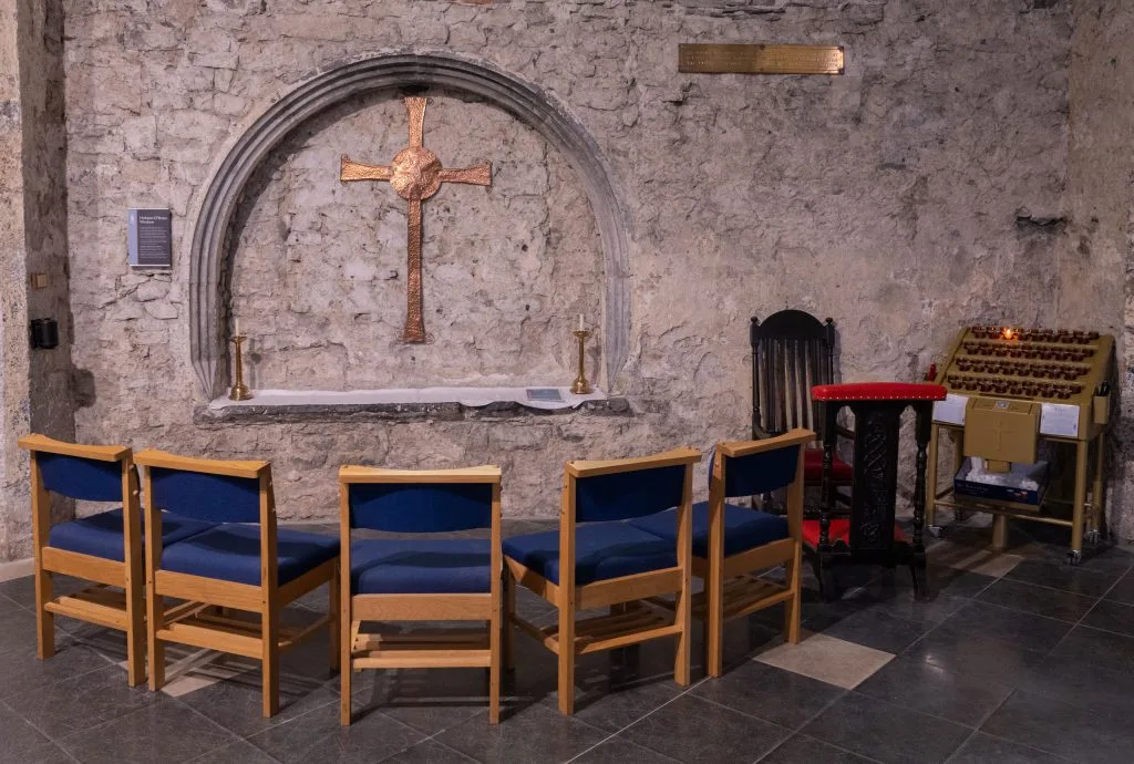 A small stone chapel interior featuring a copper cross mounted inside a stone alcove on a rough stone wall. Below the cross, two candlesticks flank a white cloth-covered ledge. In front, five wooden chairs with blue cushions are arranged in a semicircle. To the right, a wooden kneeling bench with red cushioning and a black chair stand next to a votive candle holder rack with one candle lit. The floor is tiled in large gray and cream squares.