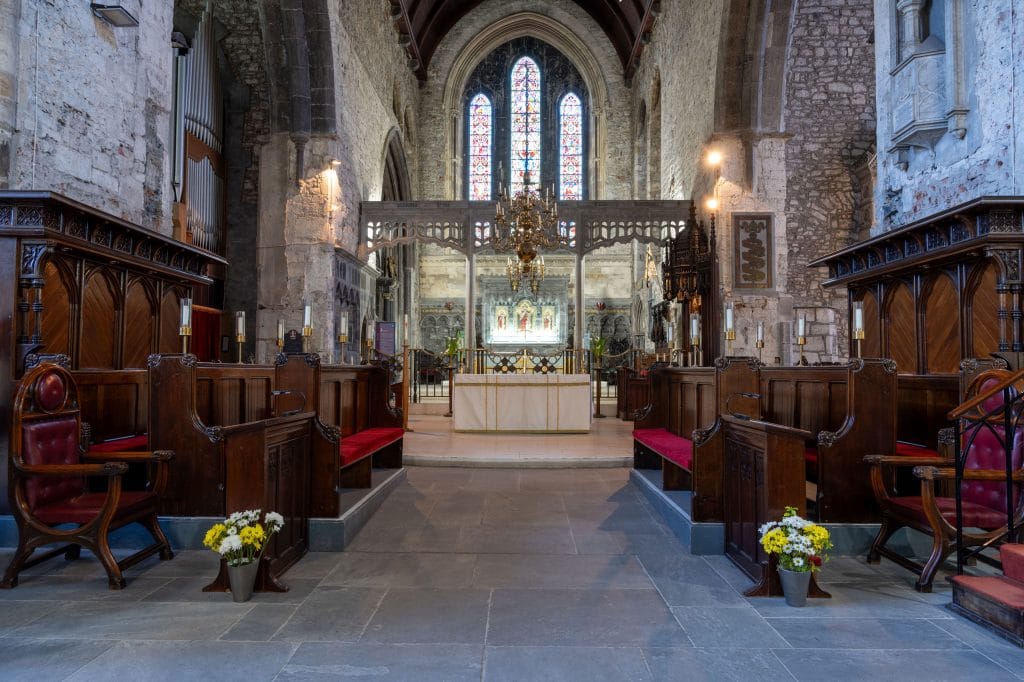 Interior of an old stone church featuring wooden pews with red cushions, an altar in the center, stained glass windows in the background, and floral arrangements on either side of the aisle.
