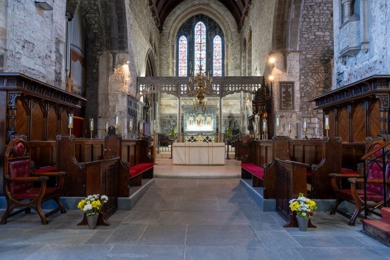 Interior of an old stone church featuring wooden pews with red cushions, an altar in the center, stained glass windows in the background, and floral arrangements on either side of the aisle.