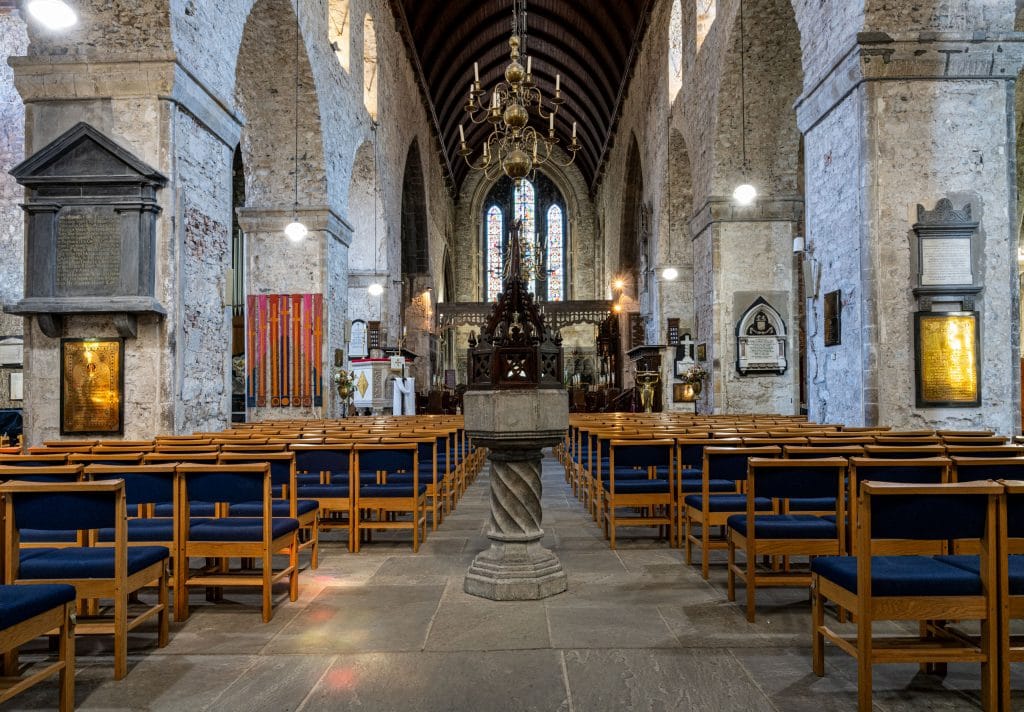 Interior of a historic church showing rows of wooden chairs with dark blue cushions, stone pillars, and arched ceilings. A decorative baptismal font stands in the center aisle, and a chandelier hangs from the ceiling. Stained glass windows illuminate the altar area in the background.