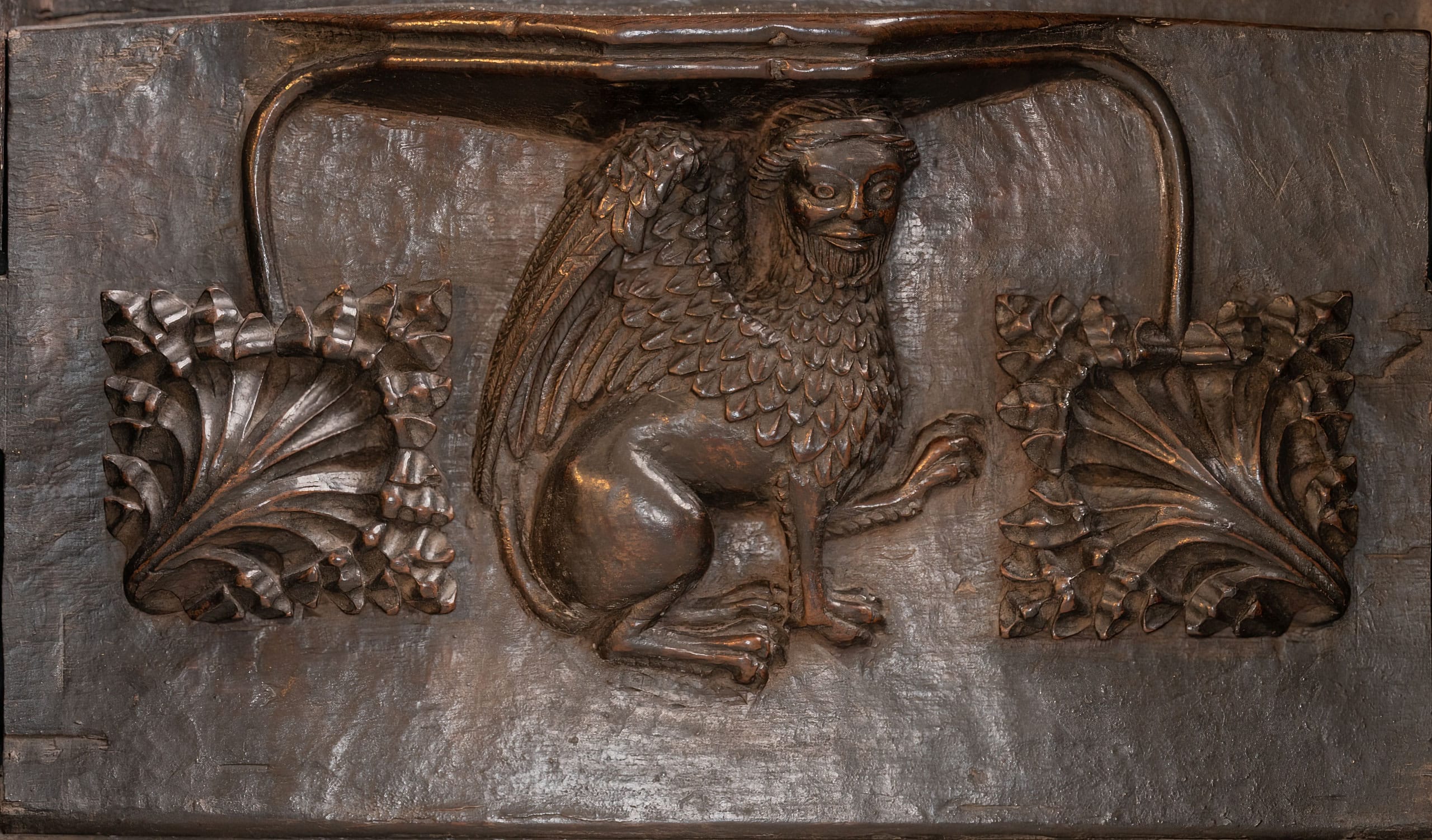 Carved wooden panel depicting a mythical griffin-like creature with a human face, detailed wings, and a raised paw, flanked by intricate leafy designs.