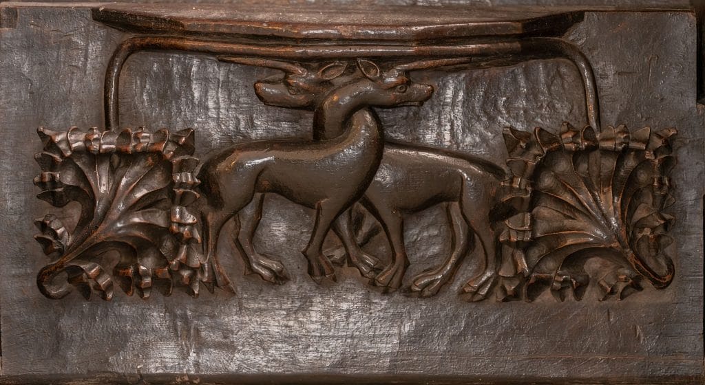 Wooden carving featuring two deer standing back to back with intertwined antlers, surrounded by ornate leaf patterns on each side.