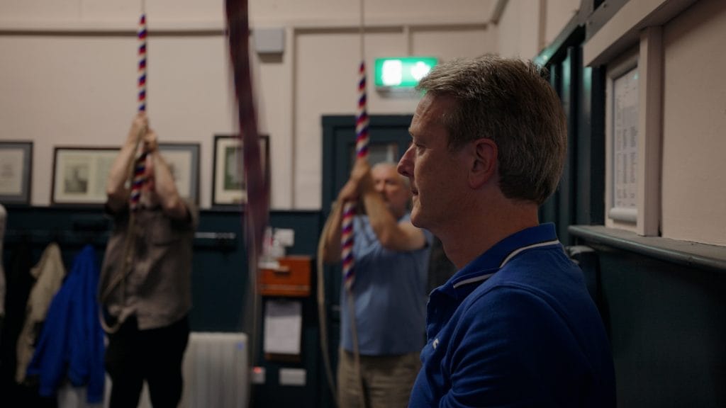 A man in a blue shirt sits in profile inside a bell-ringing room, while two other men in the background are pulling on red, white, and blue striped ropes. 