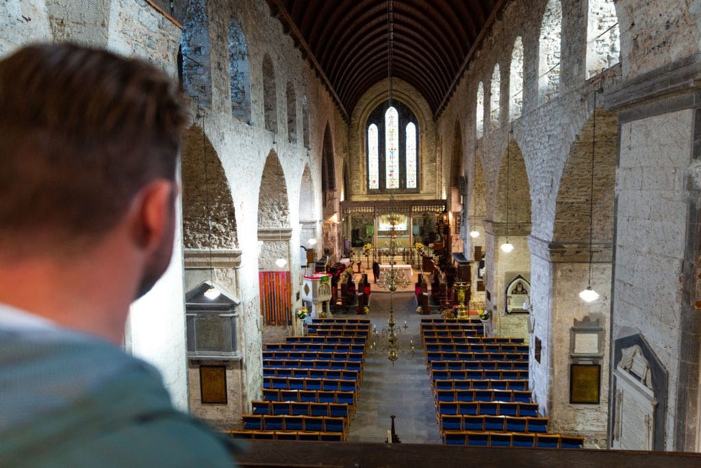 Man looking out from a balcony over the interior of a historic church with stone arches, blue cushioned wooden pews, a hanging chandelier, and stained glass windows at the far end.