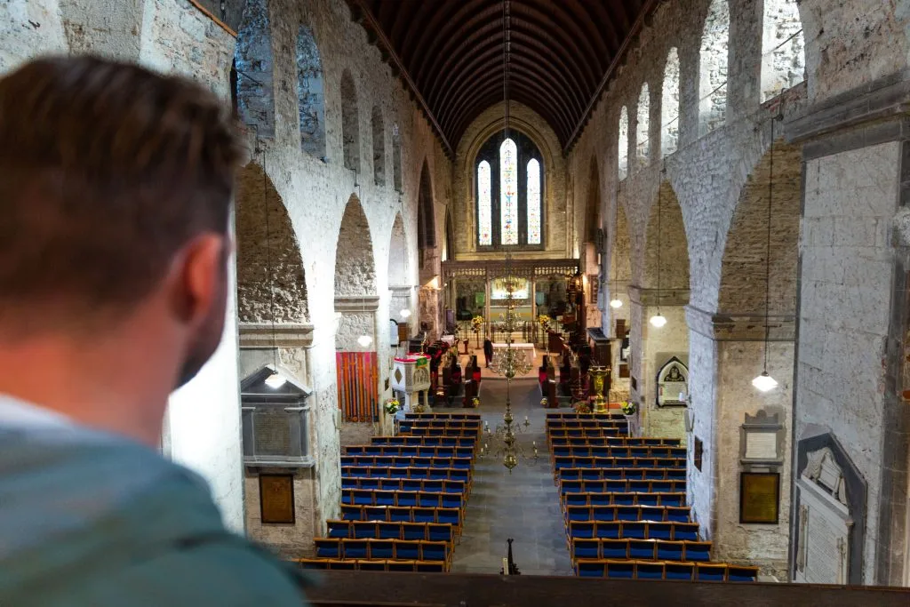 Man looking out from a balcony over the interior of a historic church with stone arches, blue cushioned wooden pews, a hanging chandelier, and stained glass windows at the far end.