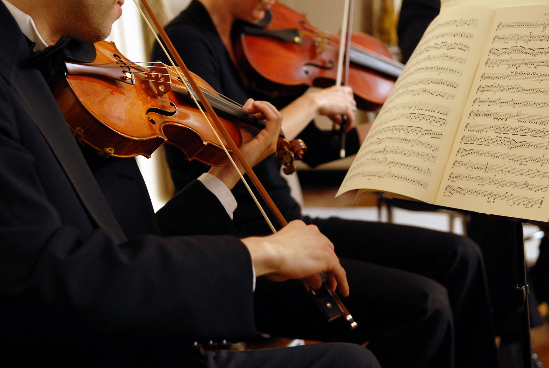 Close-up of two musicians in formal attire playing violins with sheet music on a stand in front of them.