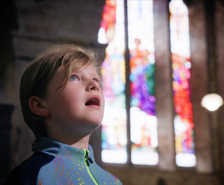 A young girl with braided hair looks upward inside a dimly lit church, with colorful stained glass windows blurred in the background.