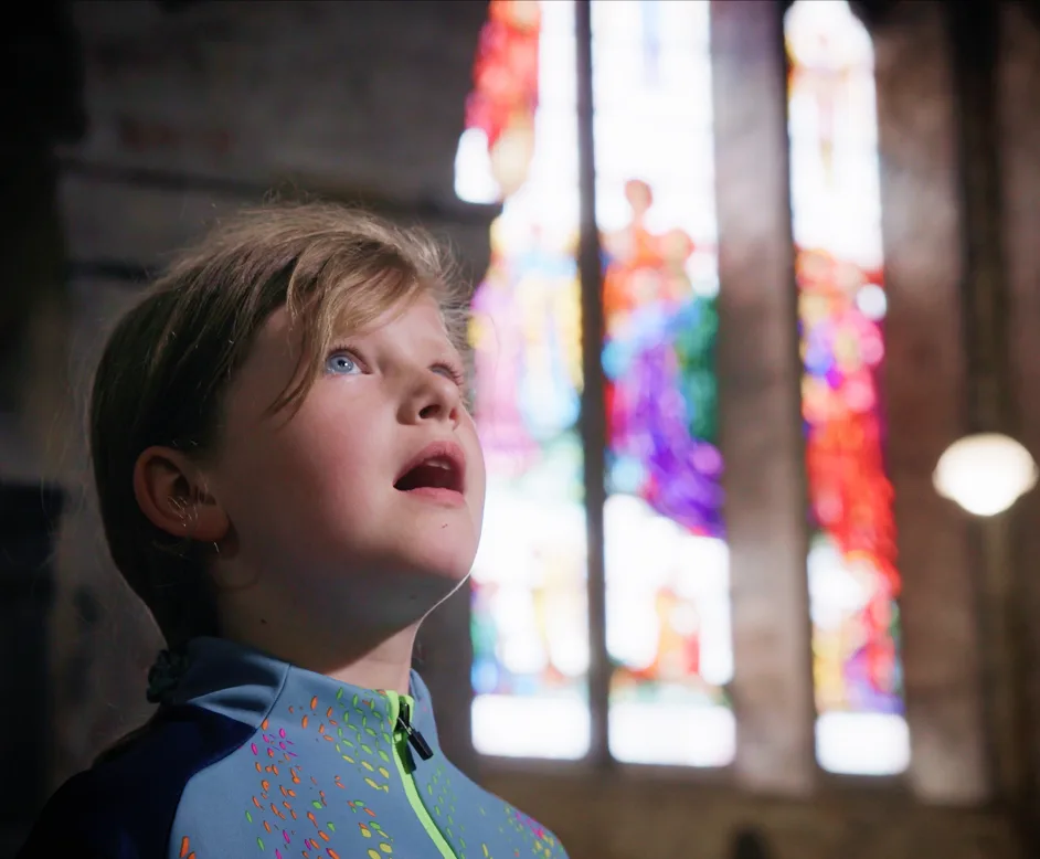 A young girl with braided hair looks upward inside a dimly lit church, with colorful stained glass windows blurred in the background.