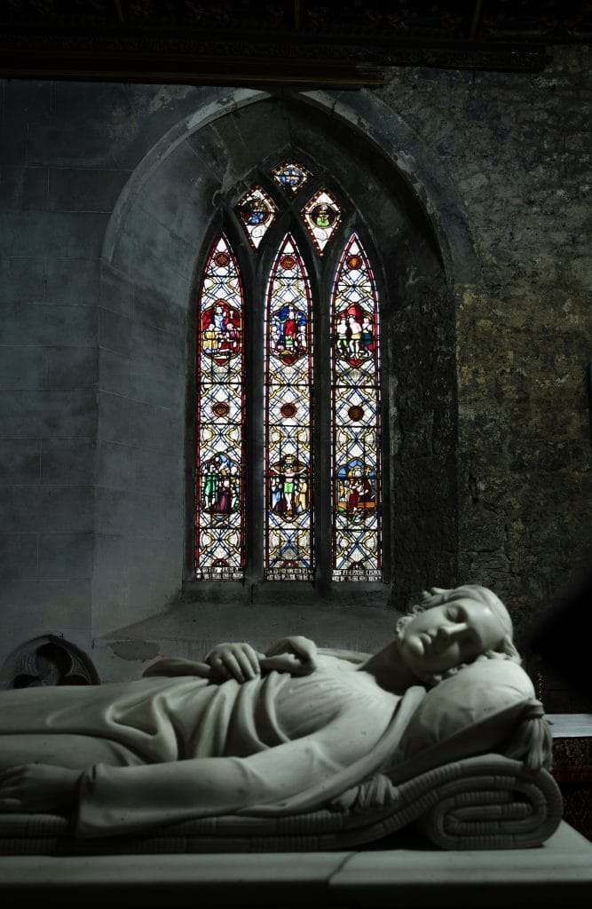A marble statue of a reclining figure with closed eyes resting on a pillow is in the foreground, set inside a stone-walled room with a large, arched stained glass window depicting religious scenes in the background.