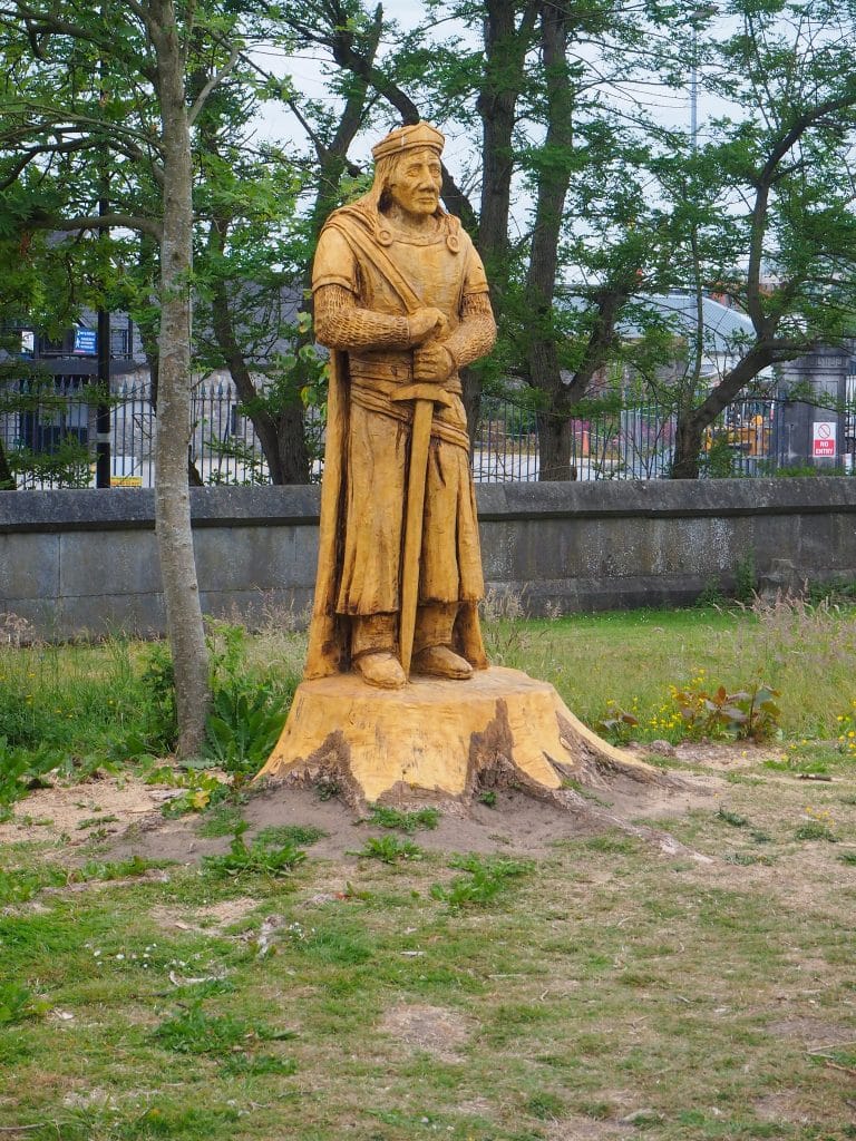 Wooden statue of a medieval knight or warrior holding a sword, carved from a tree stump, set outdoors in a grassy area with trees and a stone fence in the background.