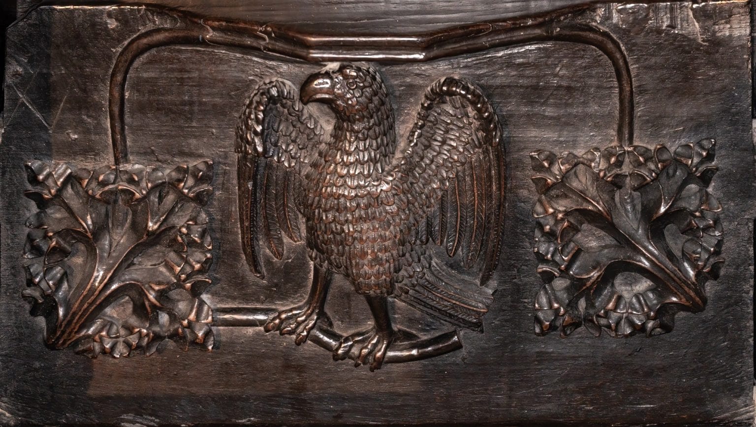 Wooden carving of a bird with spread wings perched on a branch, flanked by two symmetrical decorative leaf patterns.