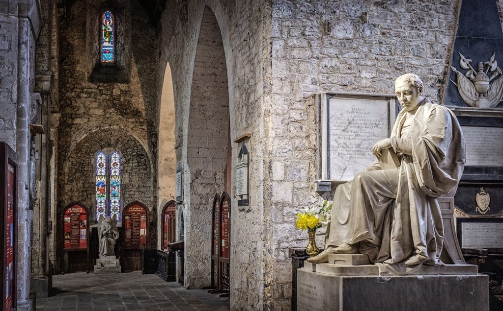 Interior view of a stone-walled cathedral featuring a seated marble statue of a robed figure on the right, with a plaque and vase of yellow flowers beside it, and stained glass windows in the background.
