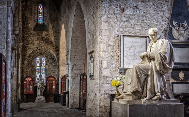 Interior view of a stone-walled cathedral featuring a seated marble statue of a robed figure on the right, with a plaque and vase of yellow flowers beside it, and stained glass windows in the background.