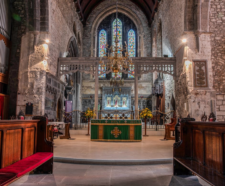 Interior of a historic church featuring wooden pews with red cushions, a green altar cloth embroidered with the word "HOLY," stone walls, stained glass windows, and a large brass chandelier hanging from a wooden vaulted ceiling.