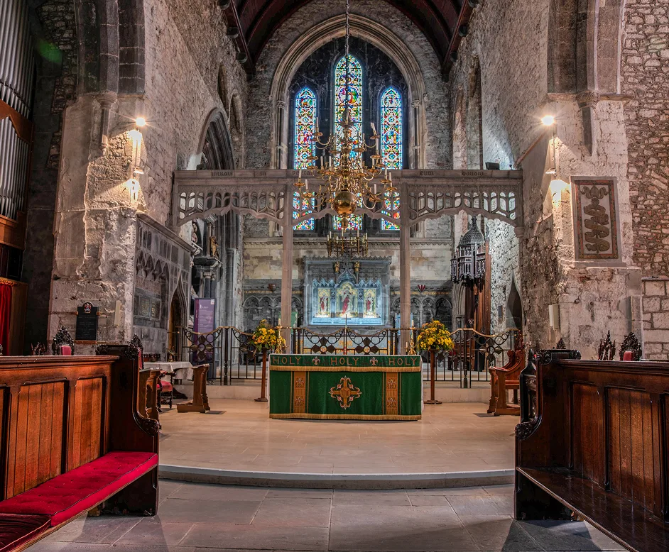 Interior of a historic church featuring wooden pews with red cushions, a green altar cloth embroidered with the word "HOLY," stone walls, stained glass windows, and a large brass chandelier hanging from a wooden vaulted ceiling.